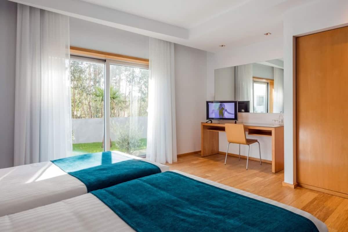 A hotel room with two beds, a desk, a TV, and a sliding glass door to the outdoors.