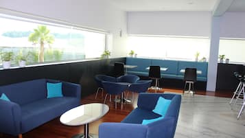A modern lounge area with blue sofas, a round table, and chairs. There are potted plants on the windowsill and a large window offering a view of palm trees.