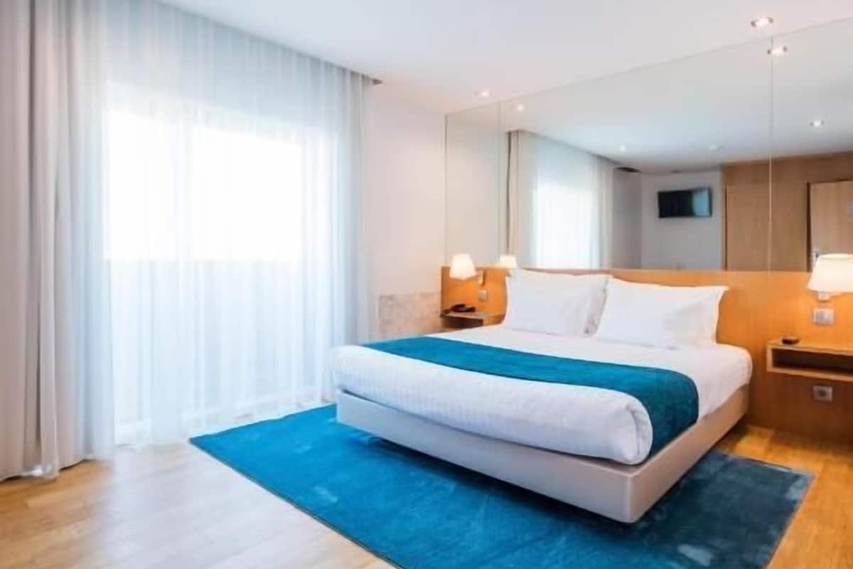A hotel room with a bed, a nightstand, a lamp, a mirror, and a window with curtains.