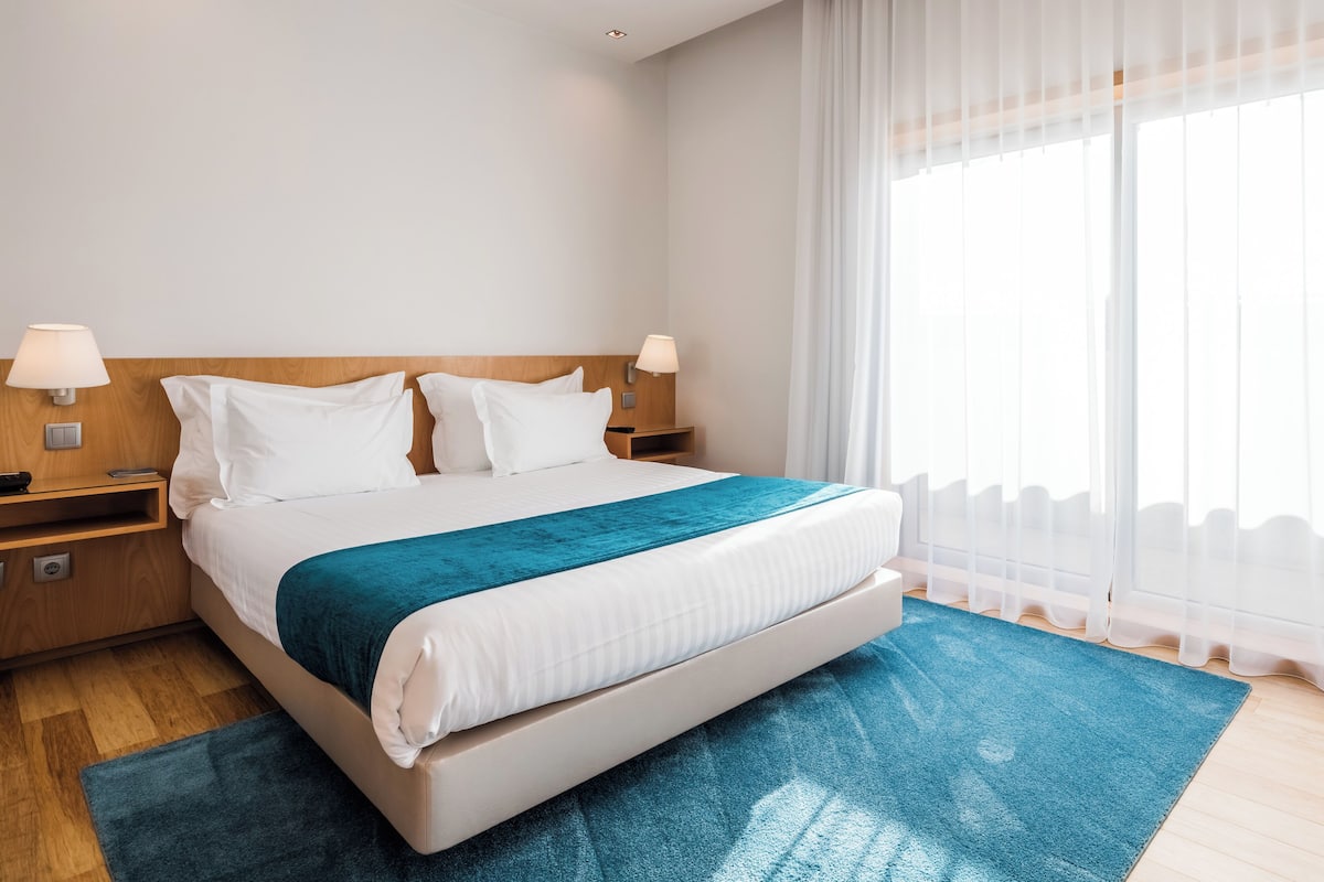 A hotel room with a large bed, two bedside tables, a wooden headboard, and a large window with sheer curtains.