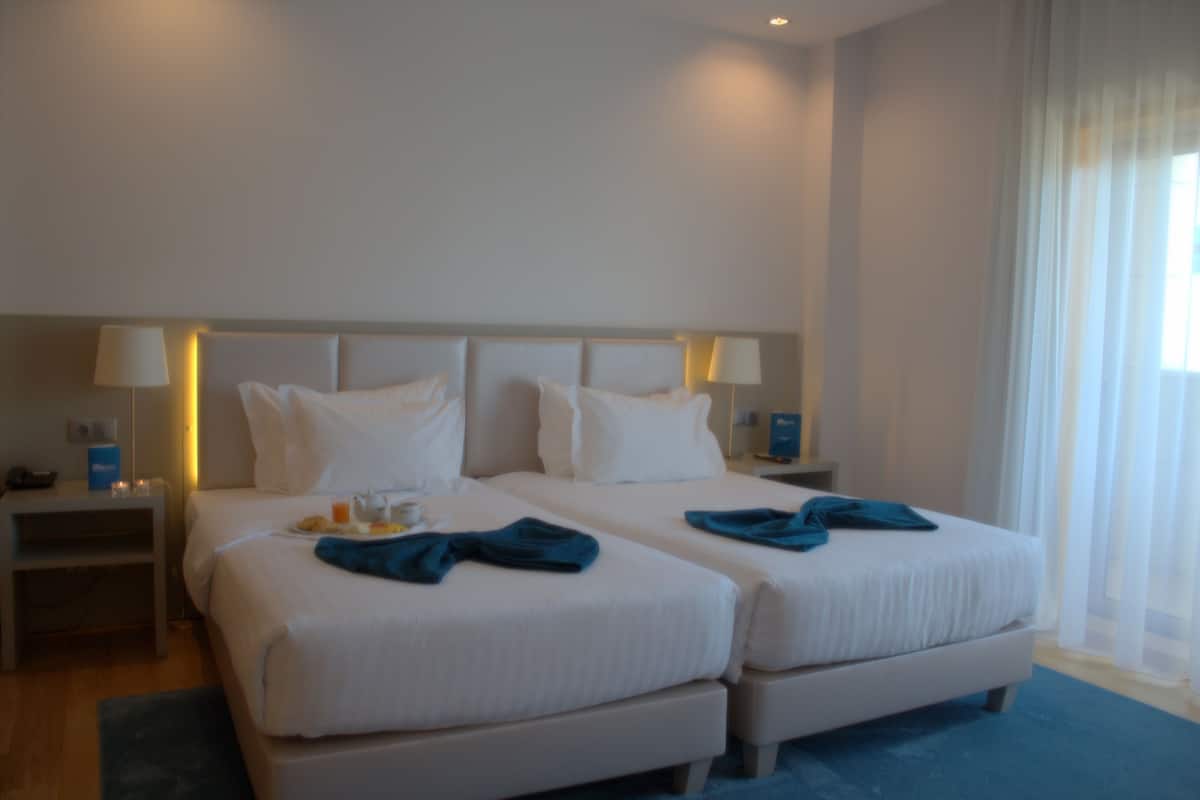A hotel room with two beds, a nightstand, a lamp, and a window with sheer curtains.