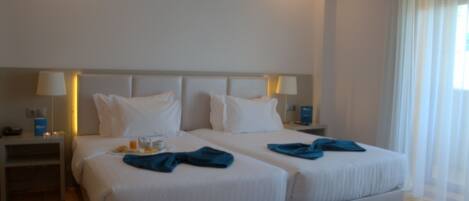 A hotel room with two beds, a nightstand, a lamp, and a window with sheer curtains.