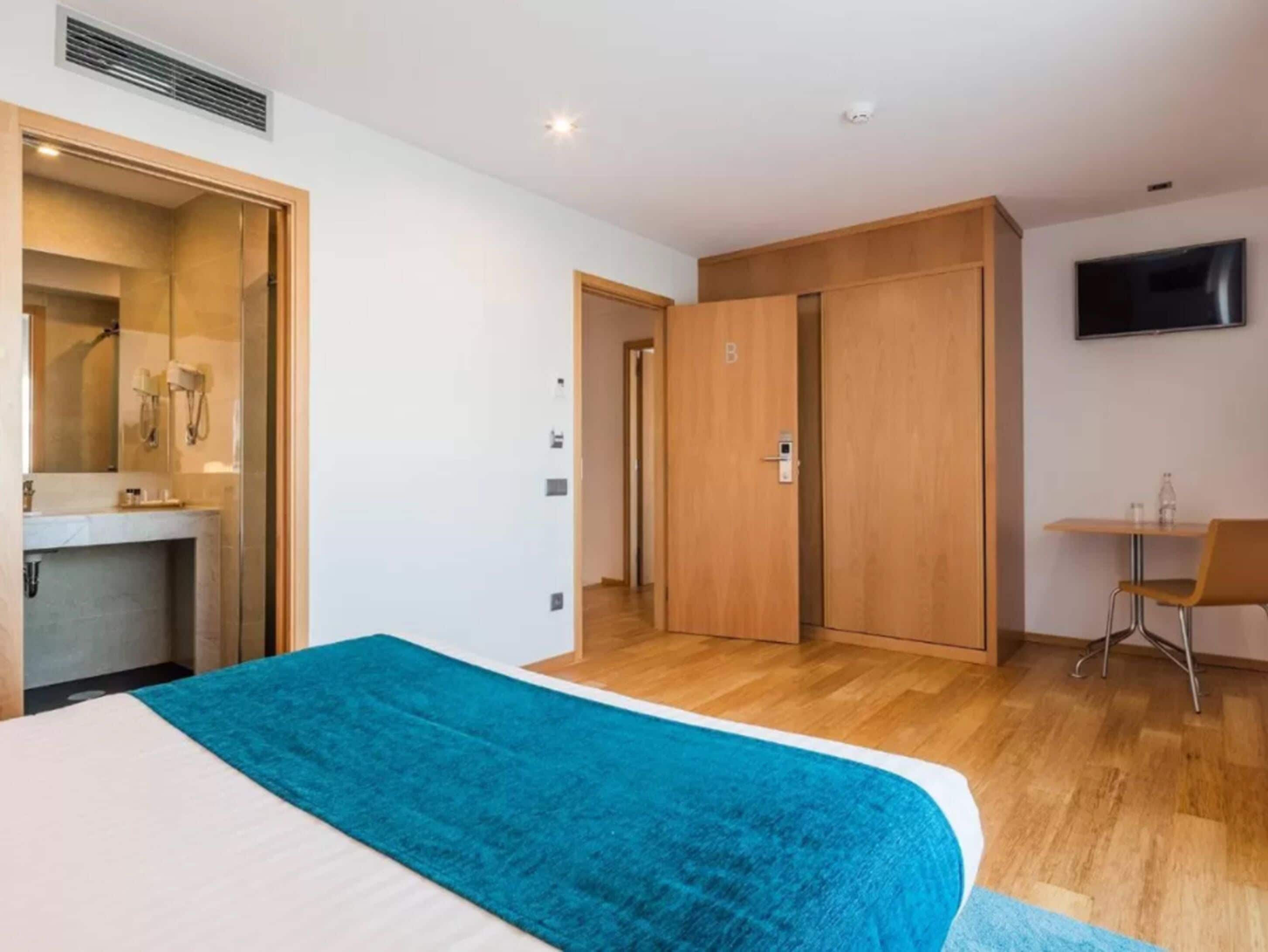 Ein Hotelzimmer mit Bett, Badezimmer mit Dusche, Schreibtisch mit Stuhl und Fernseher an der Wand.