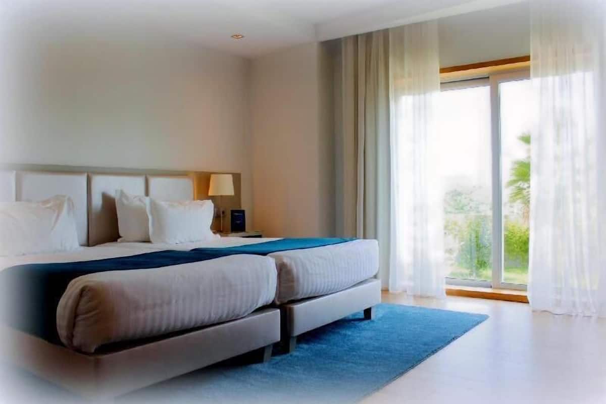 A hotel room with a bed, a nightstand, a lamp, and a view of greenery outside.