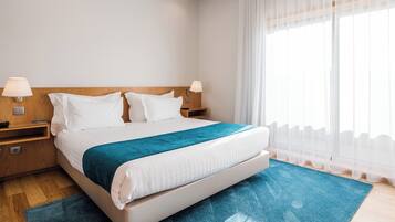A hotel room with a large bed, two bedside tables, a wooden headboard, and a large window with sheer curtains.