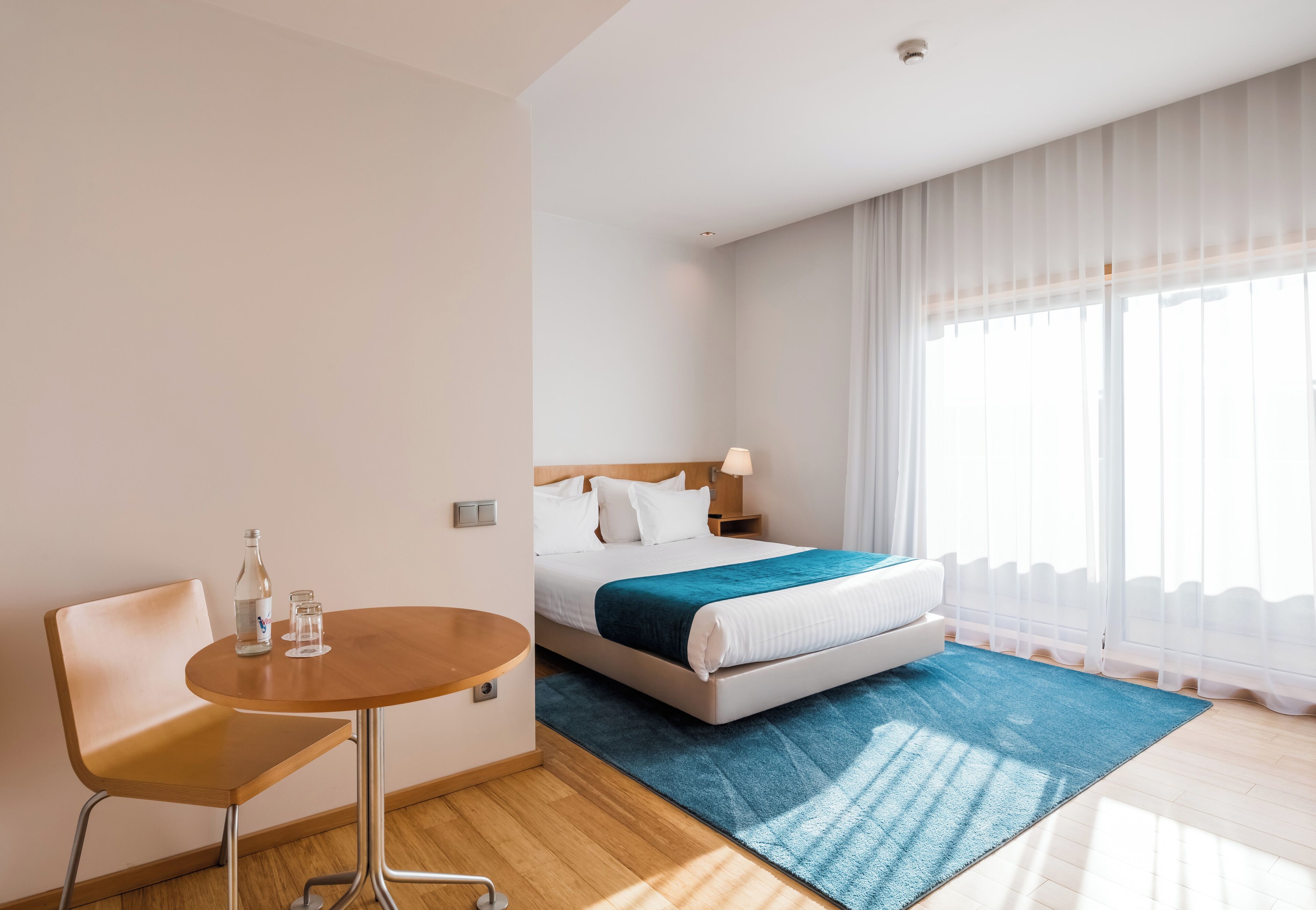 A hotel room with a bed, a bedside table with a bottle and glasses, a chair, and a large window with sheer curtains.