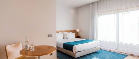A hotel room with a bed, a bedside table with a bottle and glasses, a chair, and a large window with sheer curtains.
