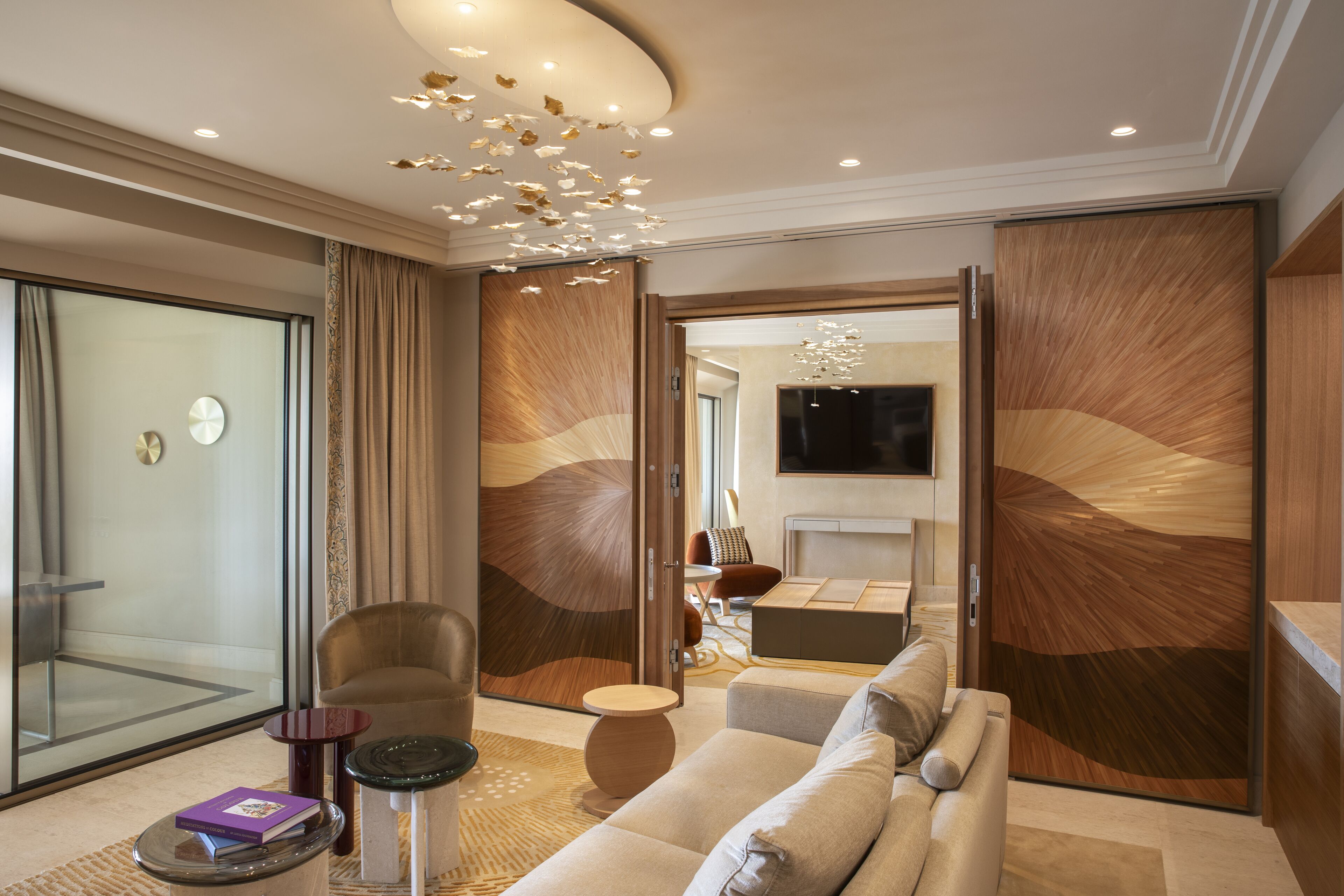 Una habitación de hotel moderna con sofá, sillón y una mesita con un libro. Hay una puerta de cristal que da a un balcón y un espejo grande en la pared.