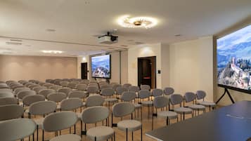 Un salón de conferencias con filas de sillas, una pantalla de proyección grande y una mesa.