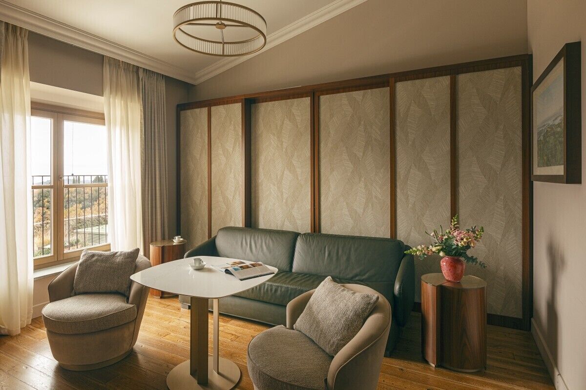 A modern living room with a leather sofa, two armchairs, a round coffee table, and a potted plant. A window with curtains provides natural light, and a framed picture is on the wall.