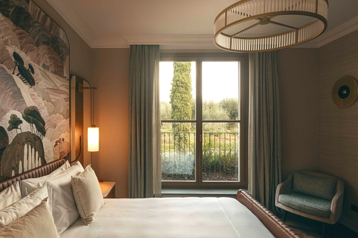 A hotel room with a large bed, a view of greenery through a window, a chair, and a wall-mounted lamp.