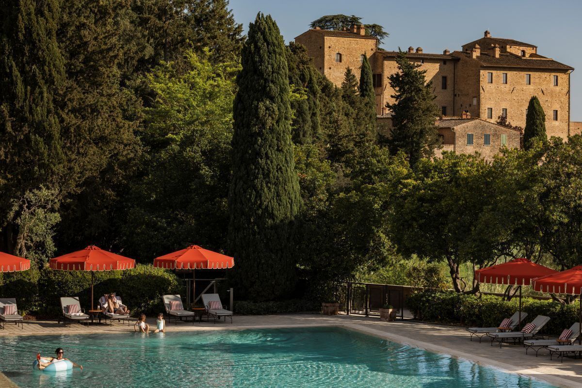 Une piscine entourée d’arbres, avec des chaises longues et des parasols, et un grand bâtiment en arrière-plan.