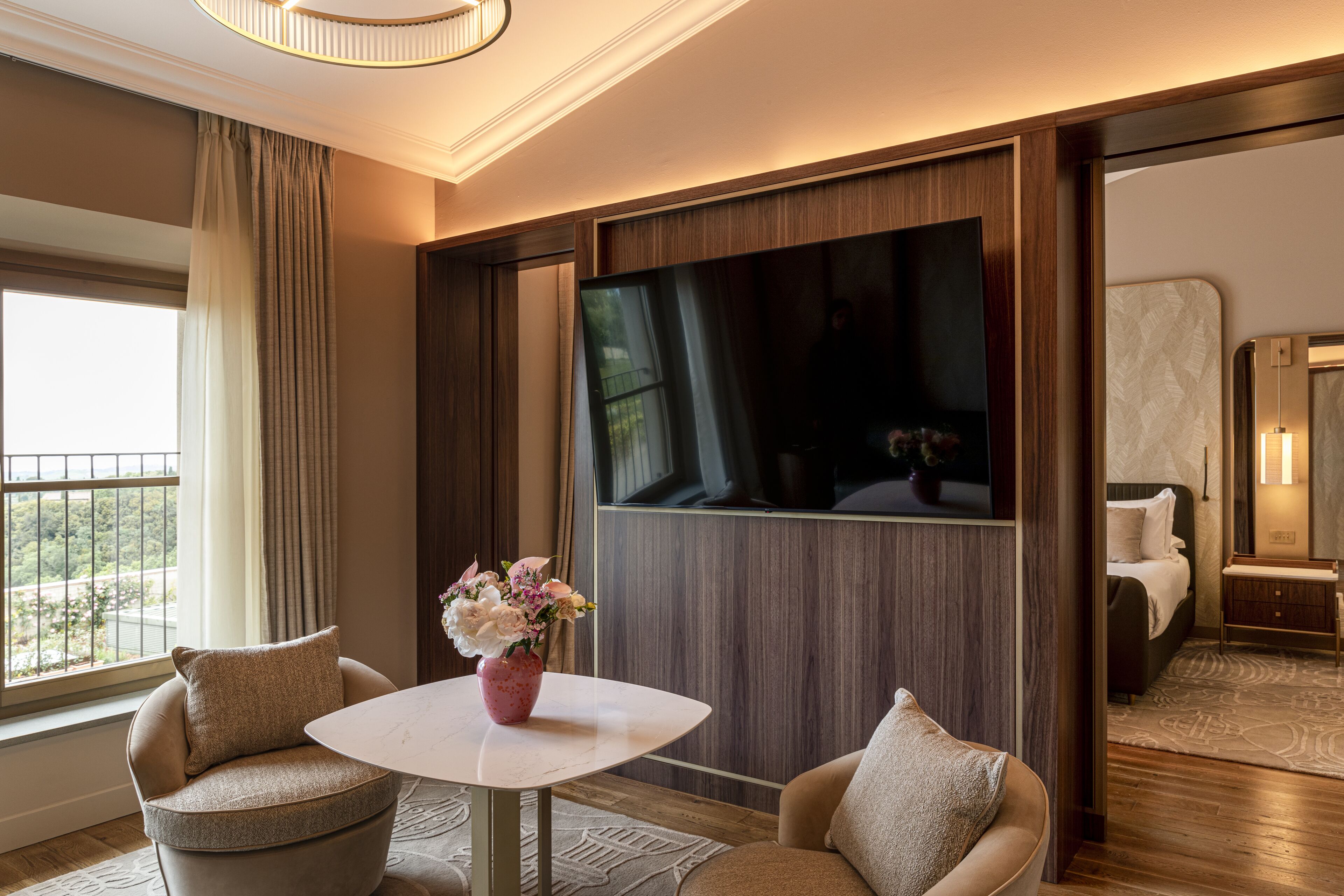 Habitación de hotel con un ventanal grande, una mesa redonda con un florero, dos sillones, una cama y un espejo.
