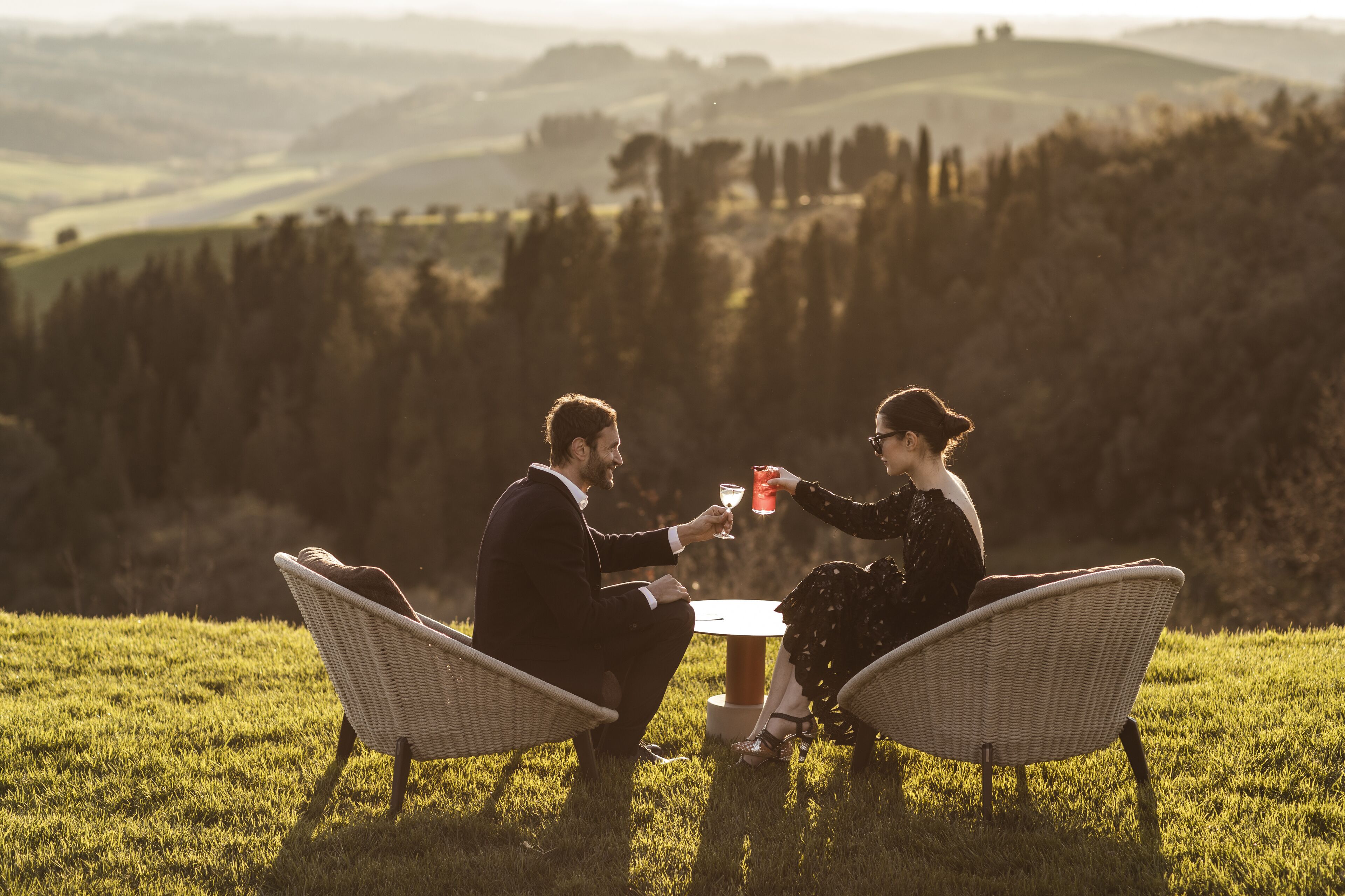 Two people sitting at a table outdoors with wicker chairs, overlooking a hilly landscape.