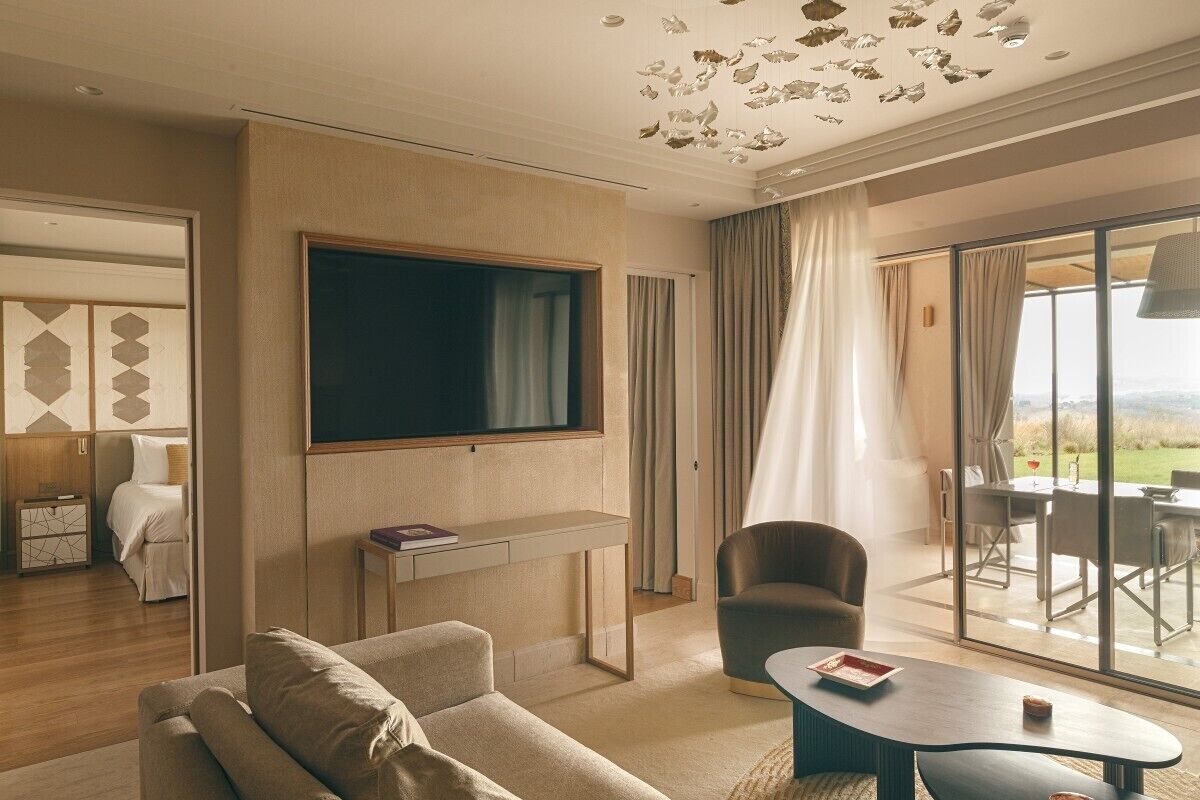 A modern hotel room with a large flat-screen TV, a comfortable sofa, a dining area with a table and chairs, and a view of the outdoors.