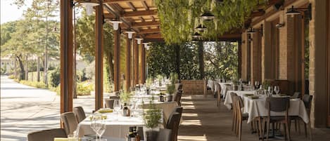An outdoor dining area with tables and chairs, covered by a wooden pergola with hanging greenery, and brick walls.