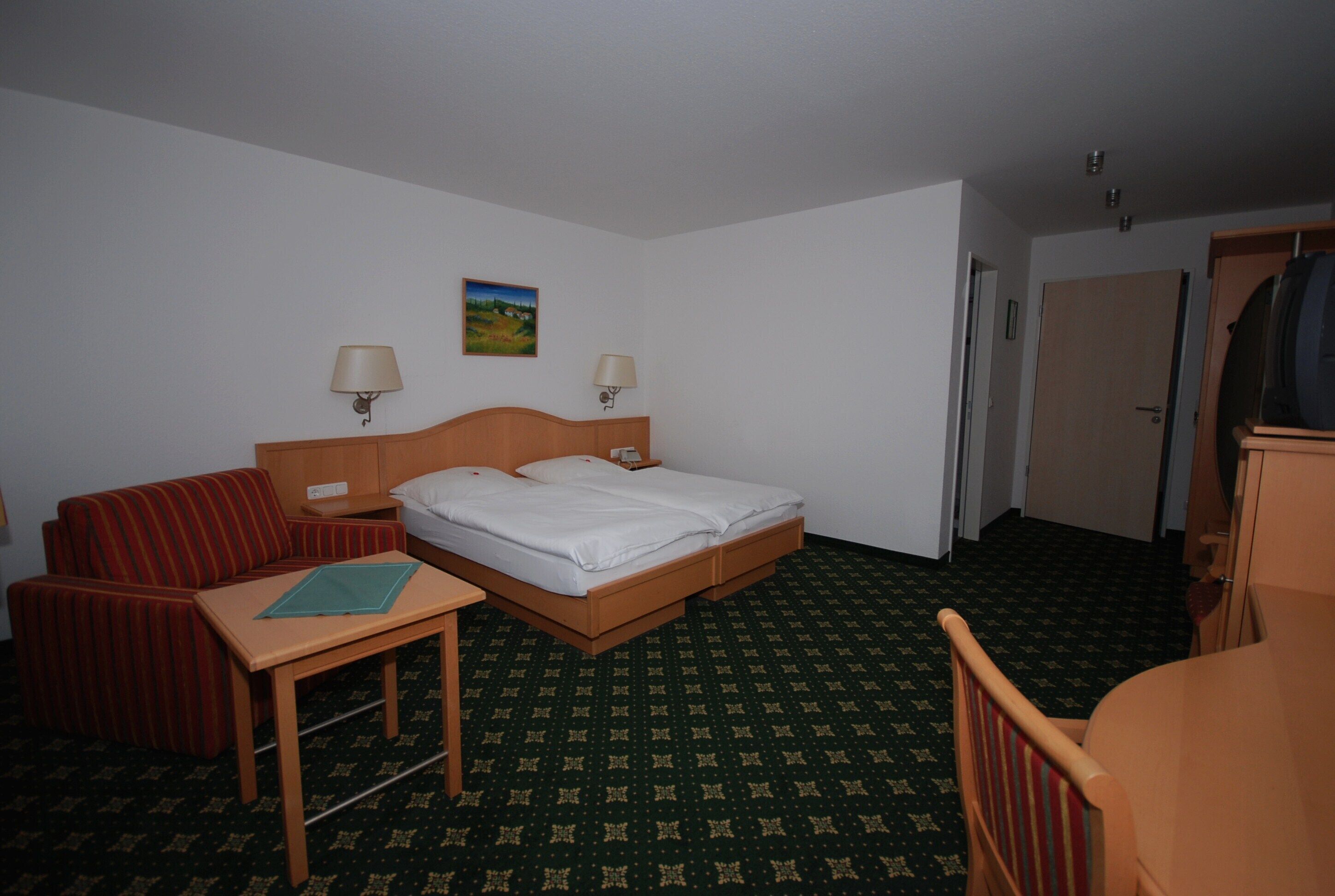 Une chambre d’hôtel avec un lit, un bureau, une chaise, une petite table, deux lampes et un tableau au mur.
