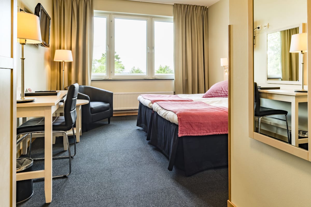 A hotel room with a desk, two chairs, a bed with a red blanket, a TV, and a window with curtains.