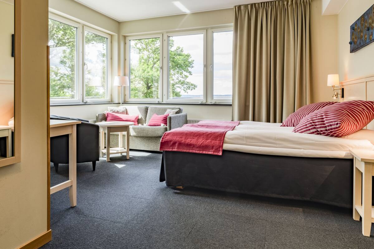 A hotel room with a bed, a desk, a chair, and large windows.