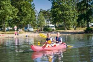 Two individuals in a red kayak on a calm body of water with trees and a sandy shore in the background. - Ringsjöstrand Hotell (Horby)
