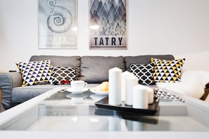 A modern living room with a grey sofa, patterned cushions, a glass coffee table with a tray, a cup, and a croissant. - EchoDom 8 Apartments Zygmunta Augusta (Kraków)