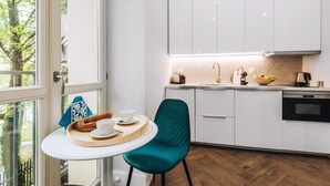 A modern kitchen with white cabinets, a teal chair, and a breakfast setup on a small table. - EchoDom 8 Apartments Zygmunta Augusta (Kraków)
