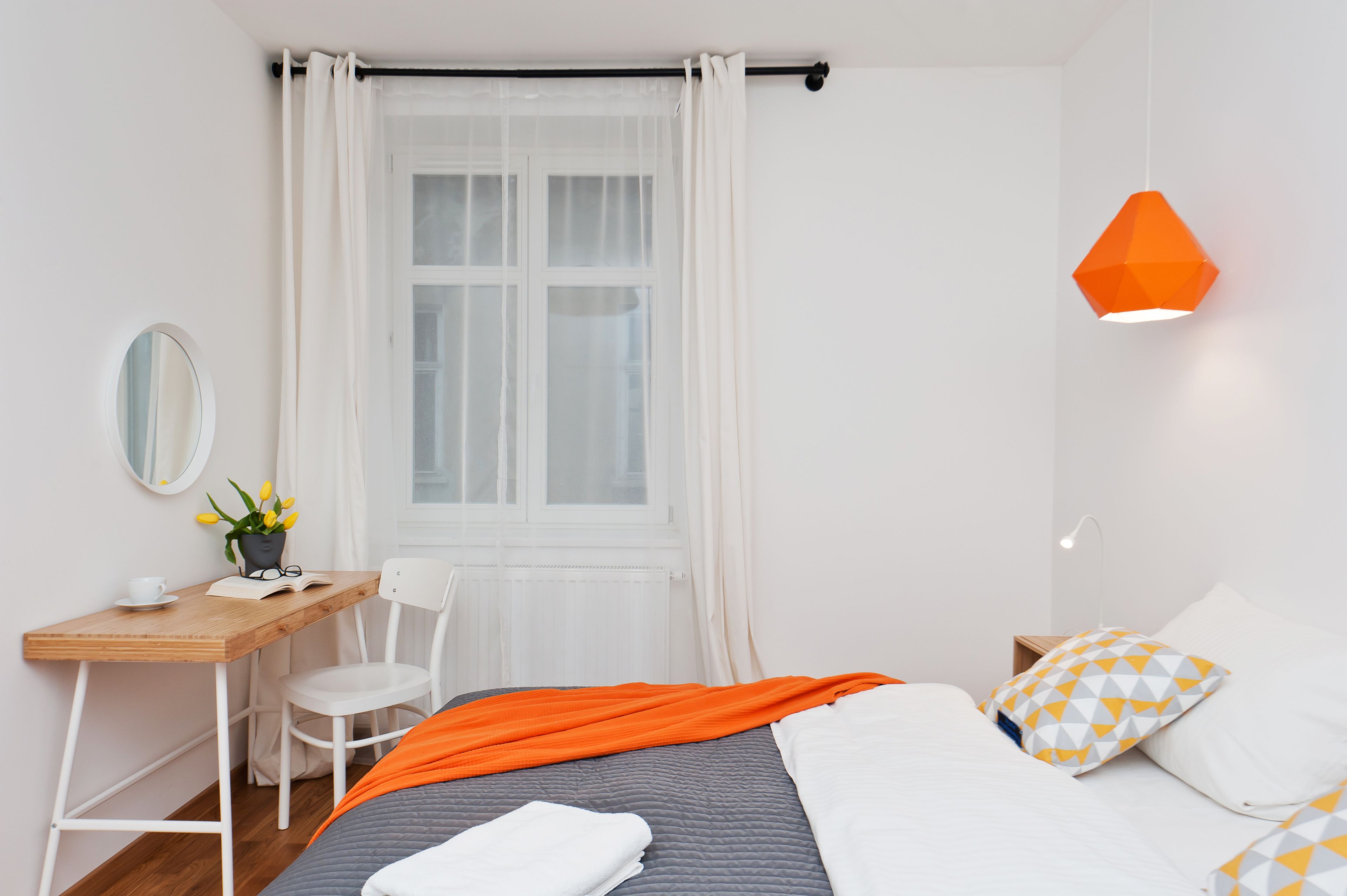Une chambre à coucher comprenant un lit, un bureau, une chaise, une fenêtre avec des rideaux et une lampe suspendue orange.