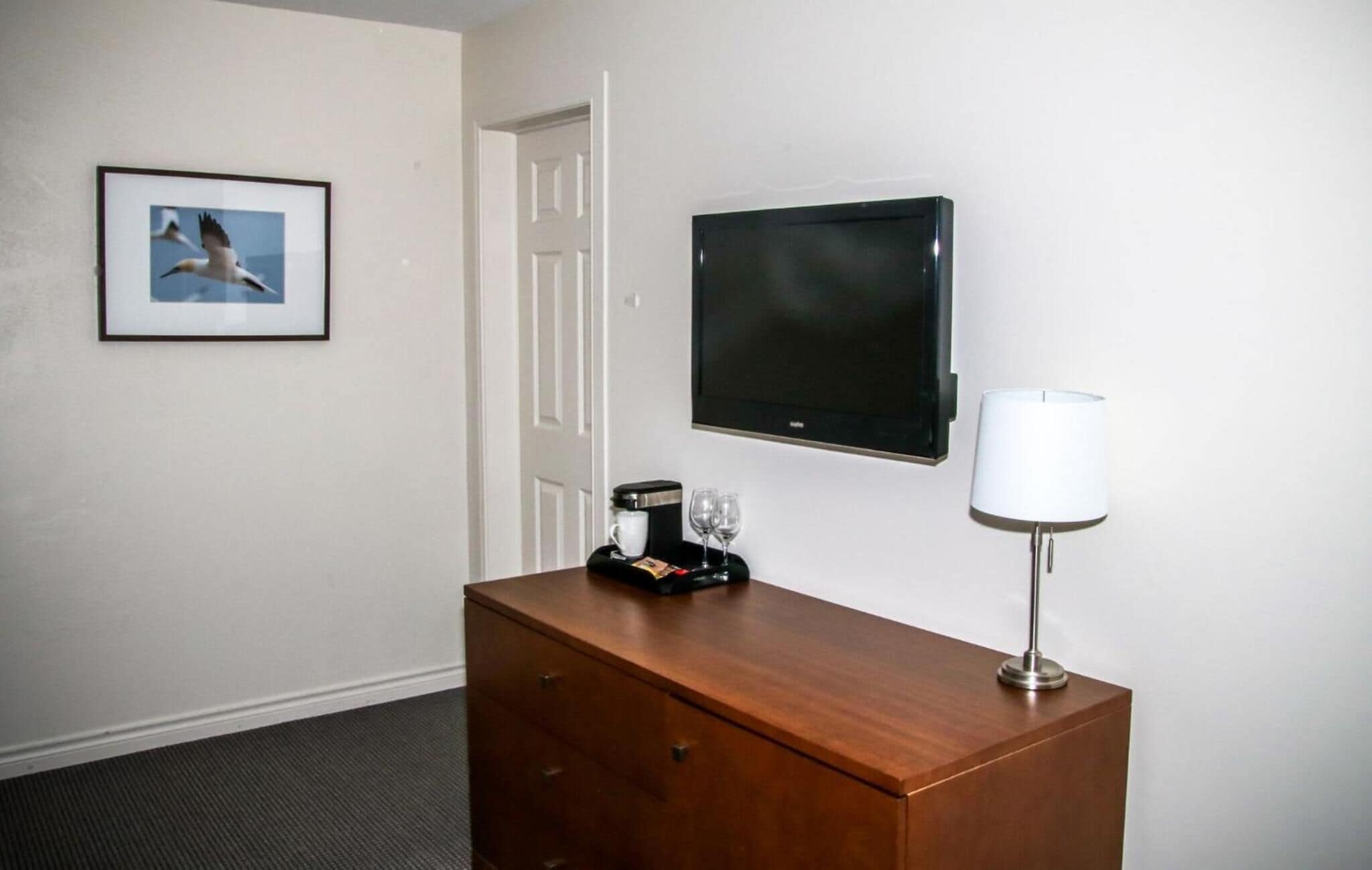 A hotel room with a wooden dresser, a flat-screen TV, a framed picture of birds, a coffee maker, and a lamp.