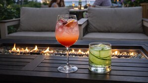 Ein Glas roter Cocktail und ein Glas grüner Cocktail auf einem Holztisch, im Hintergrund eine Feuerstelle.