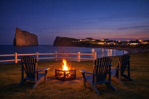 A beachfront setting with a fire pit and Adirondack chairs, overlooking a large rock formation and a lit-up coastal town.