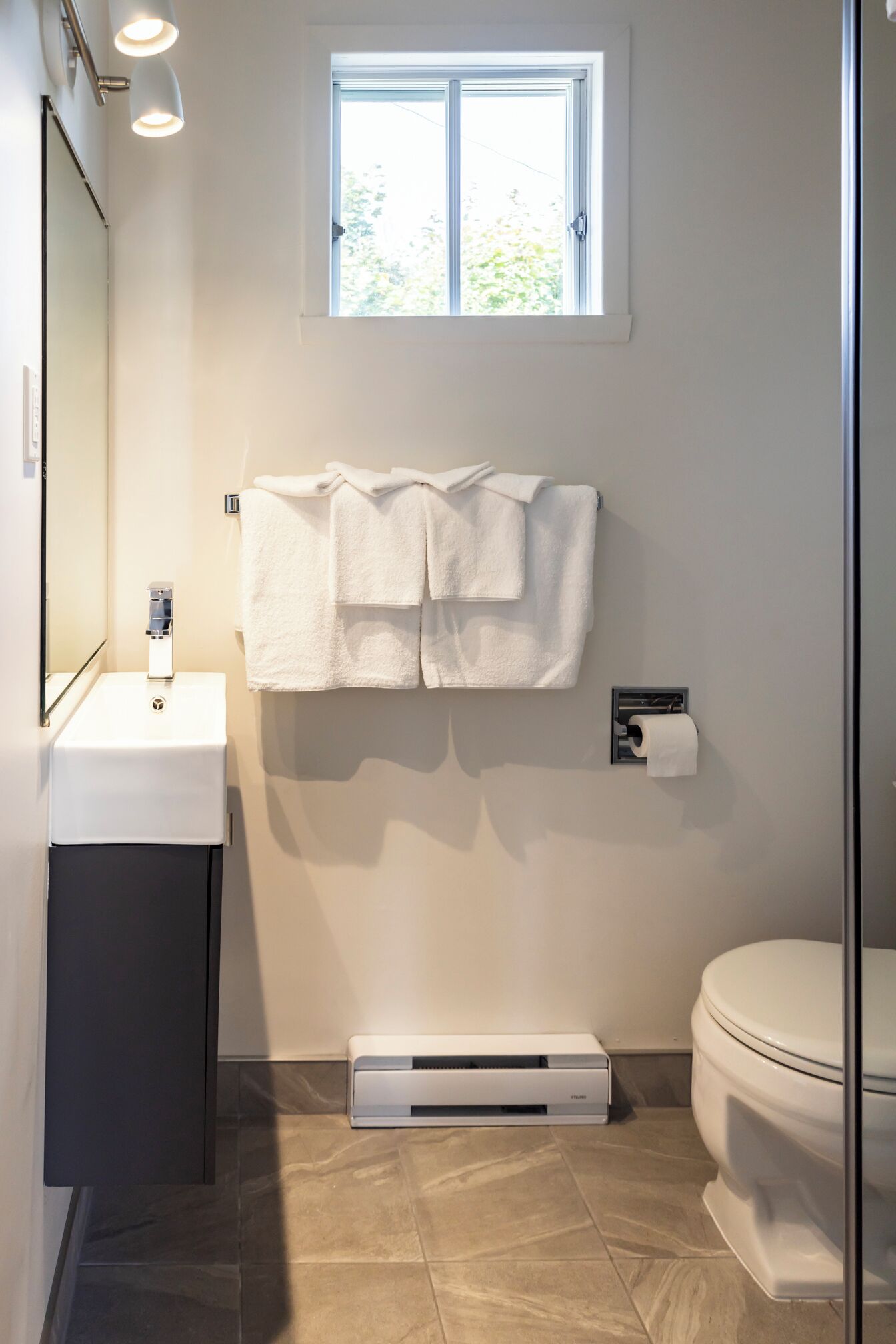 Ein modernes Badezimmer mit einem weißen Waschbecken, einem Handtuchhalter mit zwei Handtüchern, einer Toilette und einem Fenster über dem Waschbecken.