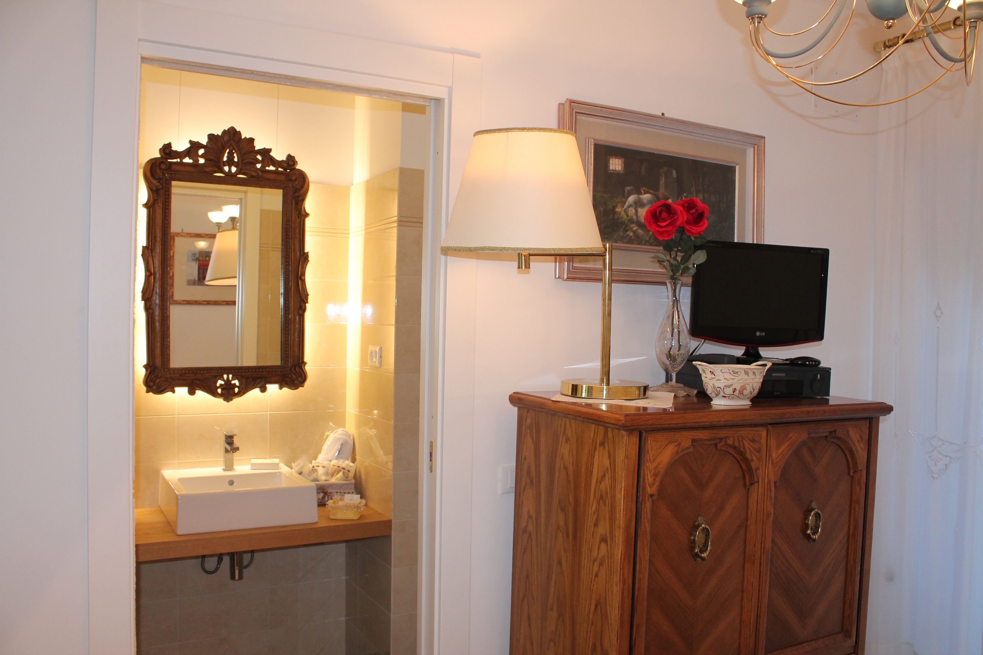 A bathroom with a wooden vanity, a mirror, a sink, a lamp, and a vase with red flowers.