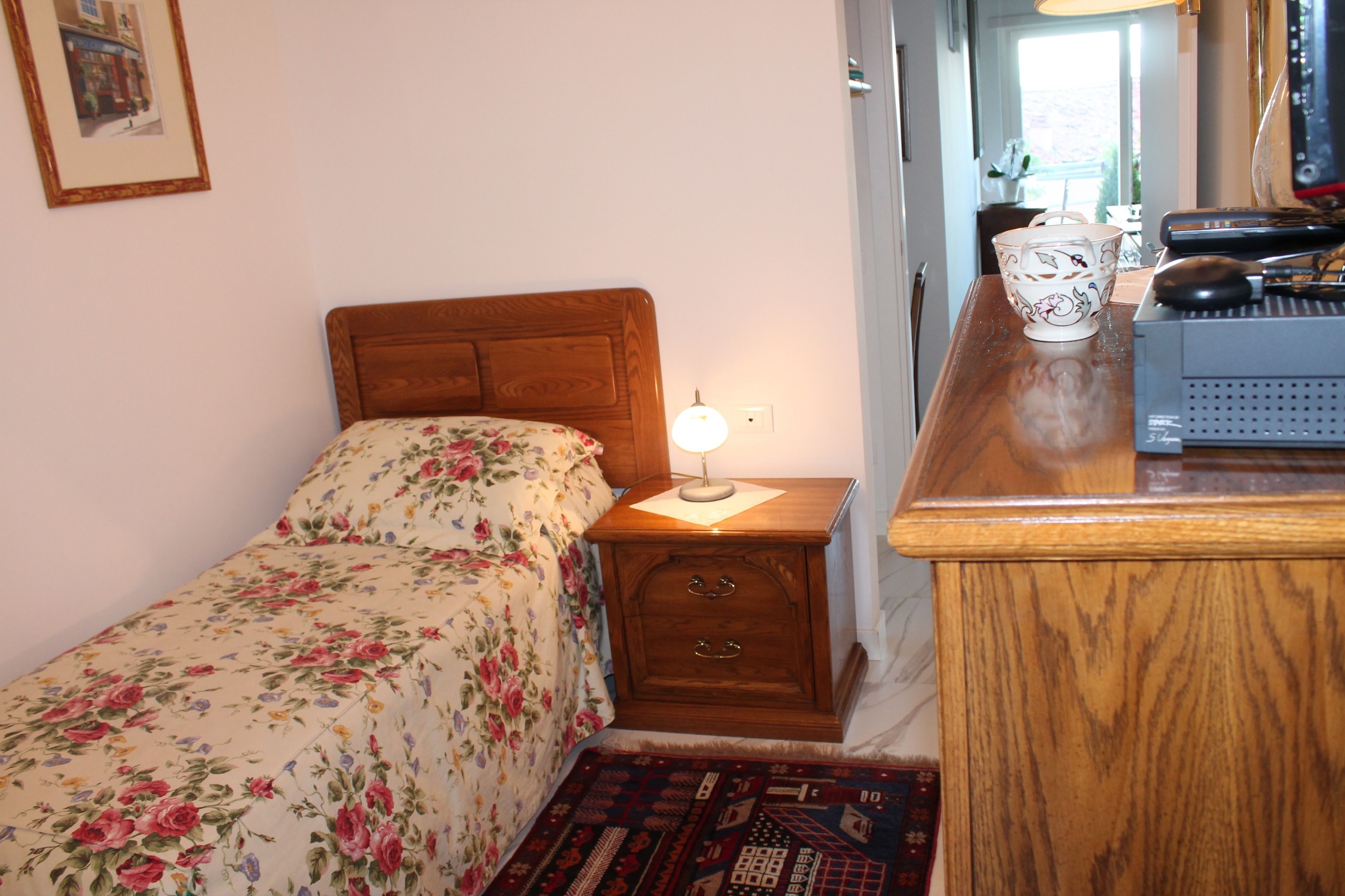 A bedroom with a wooden headboard, a bedside table with a lamp, a small desk with a computer, and a floral bedspread.