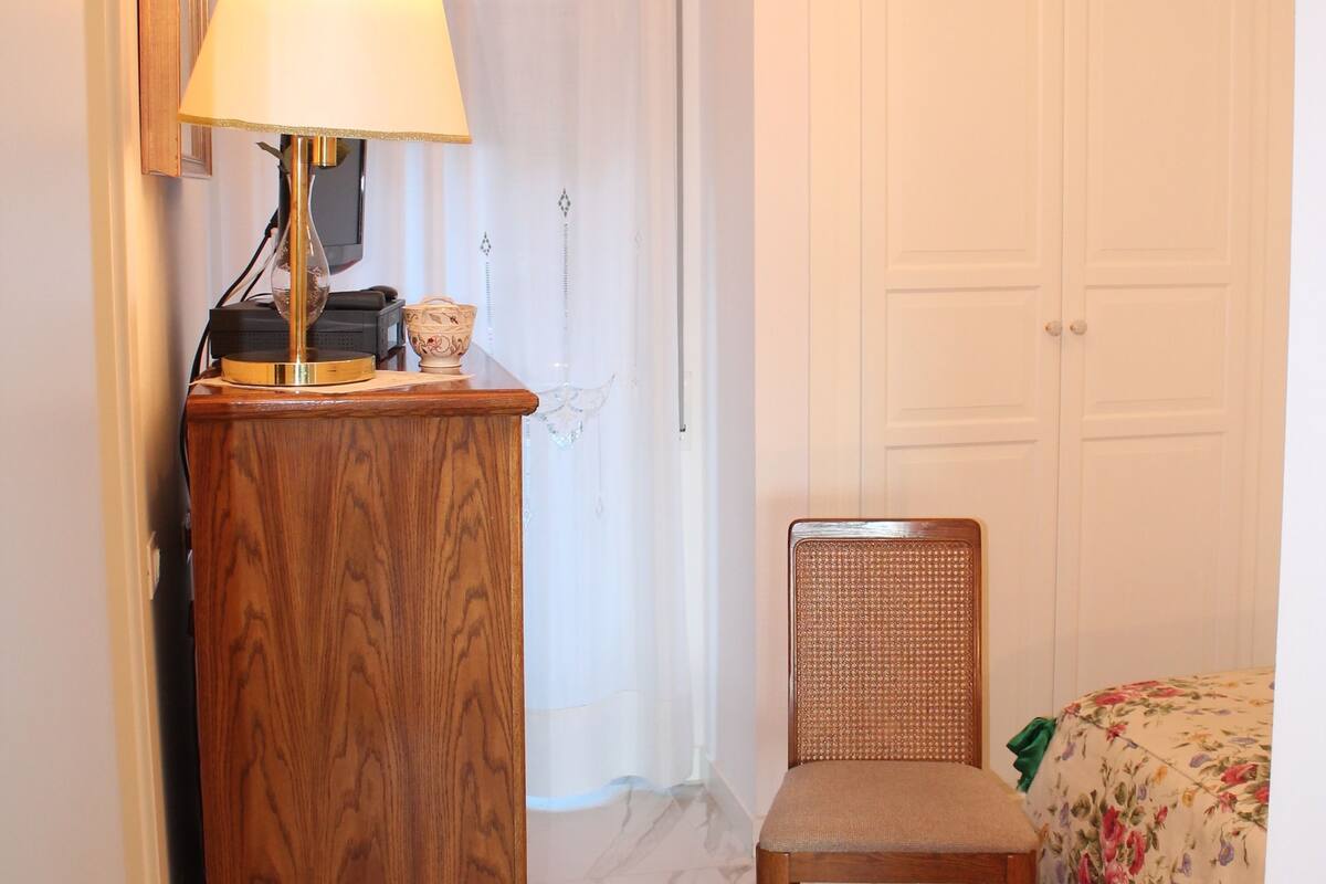 A room with a wooden desk, a chair, a bed with floral bedding, a chandelier, and a patterned rug.