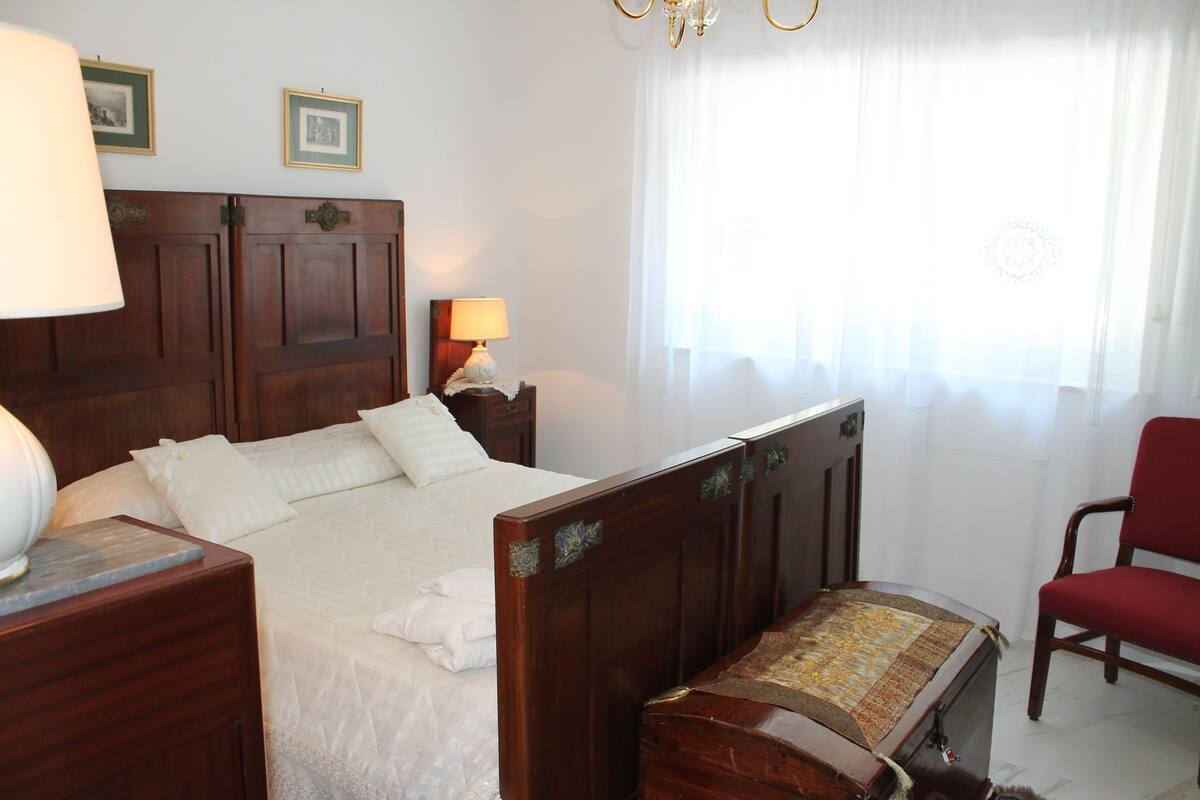 A bedroom with a wooden bed, a nightstand with a lamp, a chair, and a window with sheer curtains.