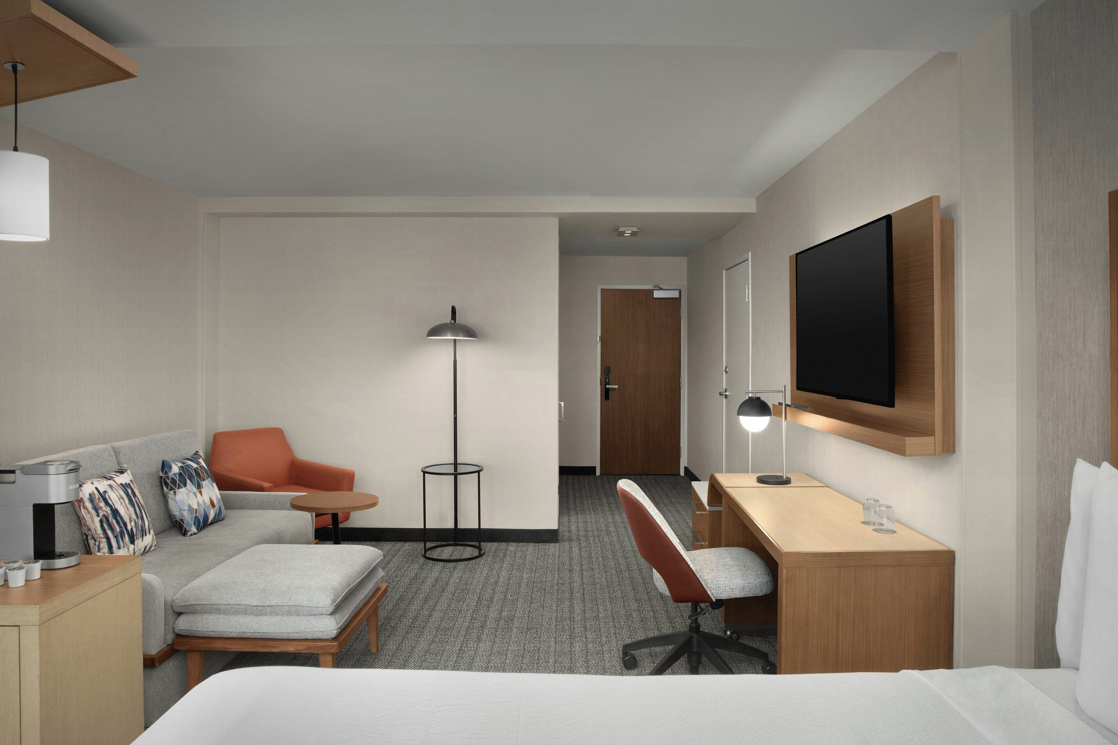 A hotel room with a bed, a desk, a chair, a sofa, a coffee machine, a lamp, and a TV.