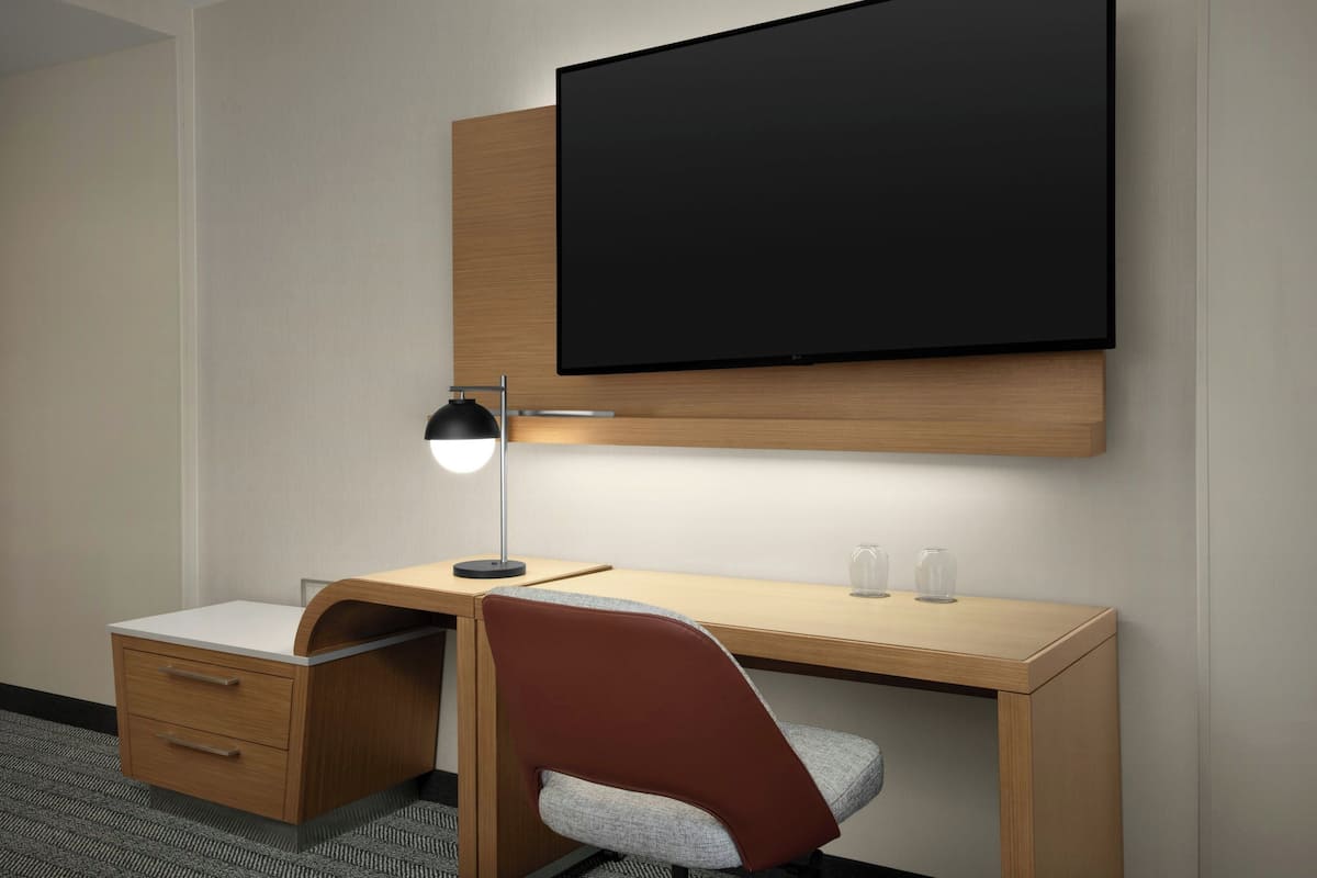 Une chambre d’hôtel moderne avec un bureau en bois, un téléviseur à écran plat fixé au-dessus, une lampe de bureau, une chaise et une petite table de chevet avec des tiroirs.