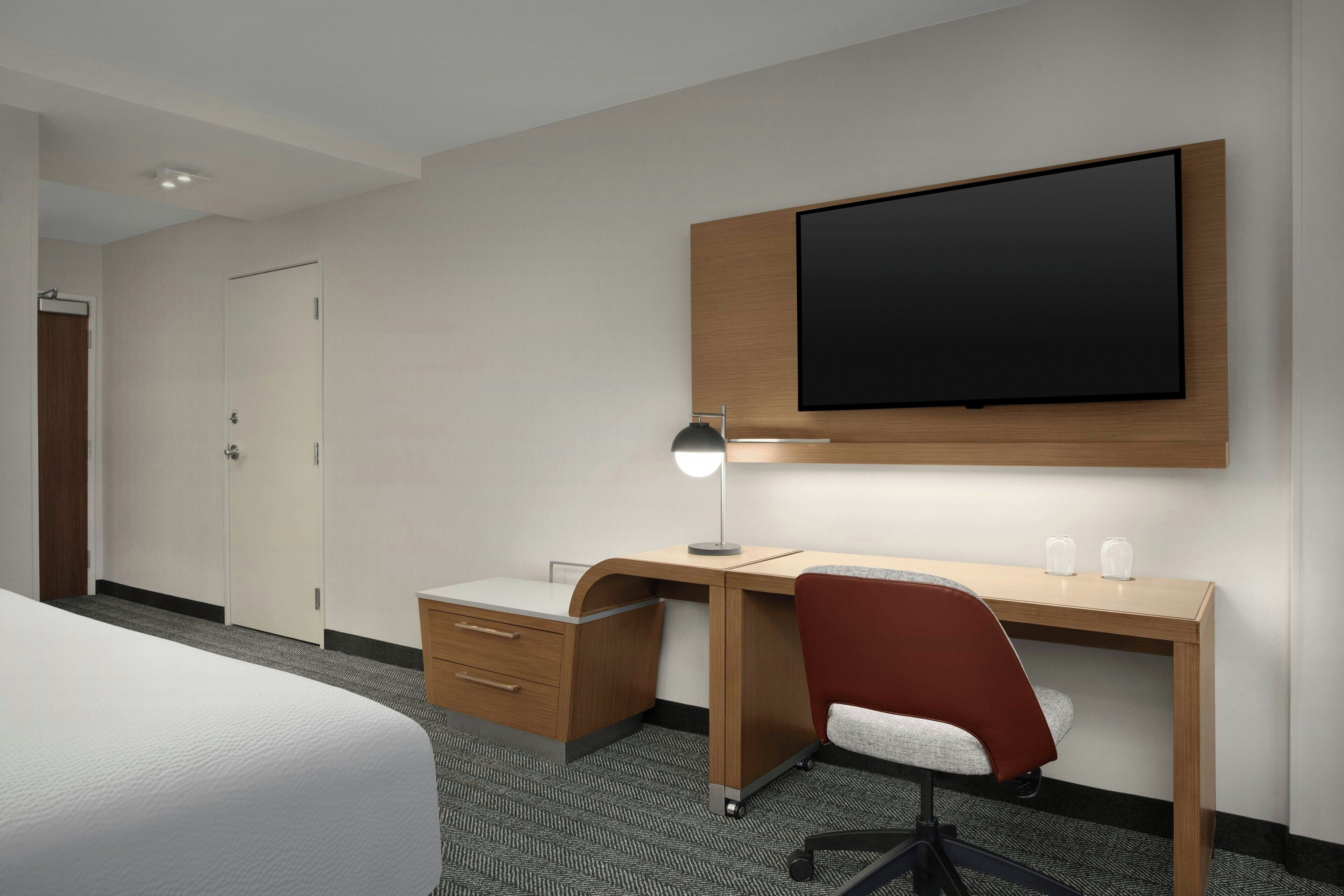 Una camera d'albergo con un letto, una scrivania con una sedia, una TV a schermo piatto montata a parete e una lampada da tavolo.