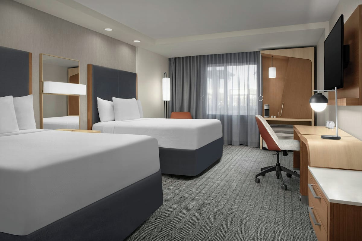 Une chambre d’hôtel avec deux lits, un bureau, une chaise, une télévision et une grande fenêtre avec des rideaux.