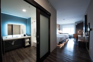 A modern hotel room with a large bed, a separate bathroom with a mirror, a desk, and a chair.