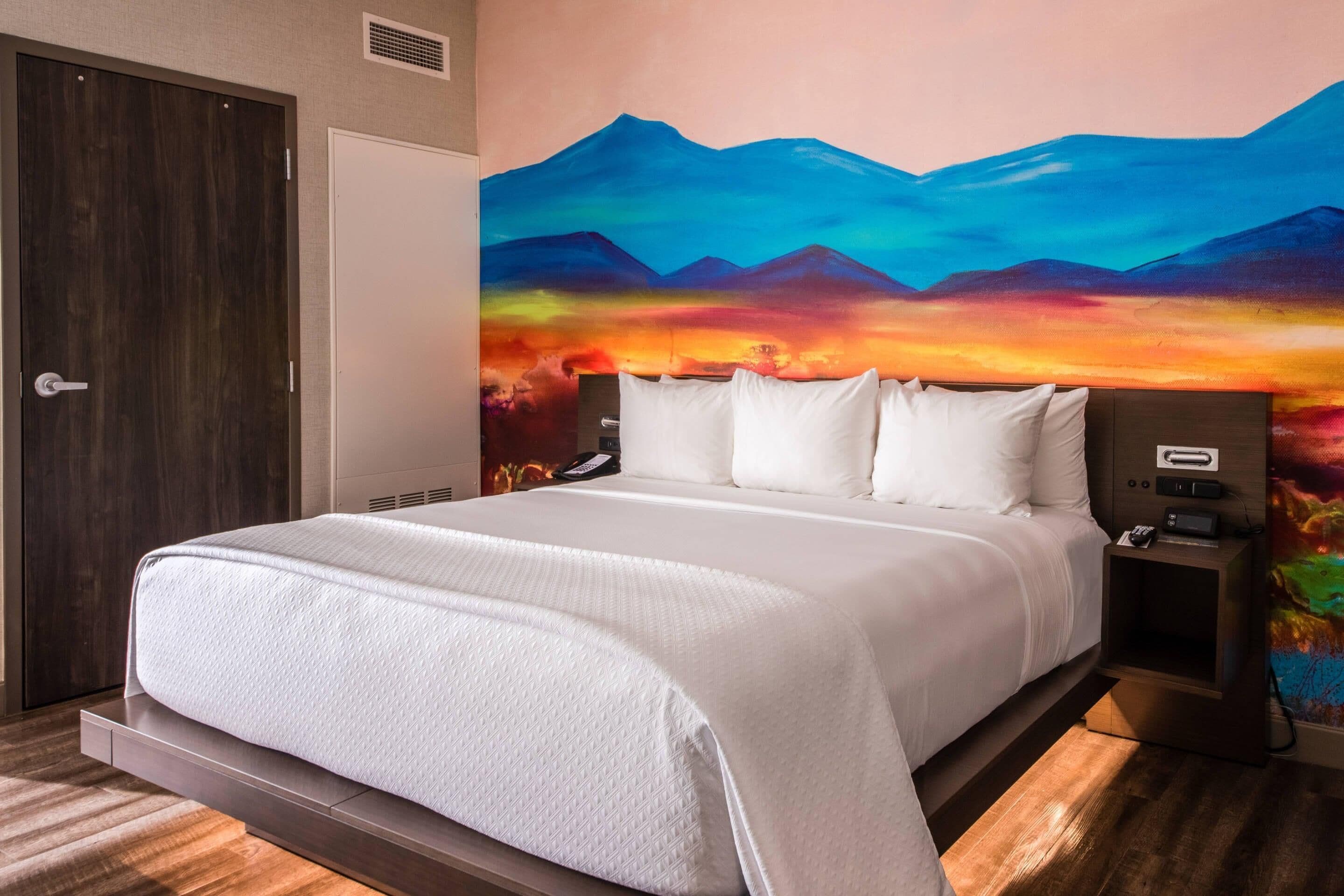 Habitación de hotel con una cama grande, una mesita de noche, un teléfono y un cuadro de un paisaje montañoso en la pared.