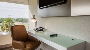 A hotel room with a desk, chair, telephone, remote control, and a TV mounted on the wall.