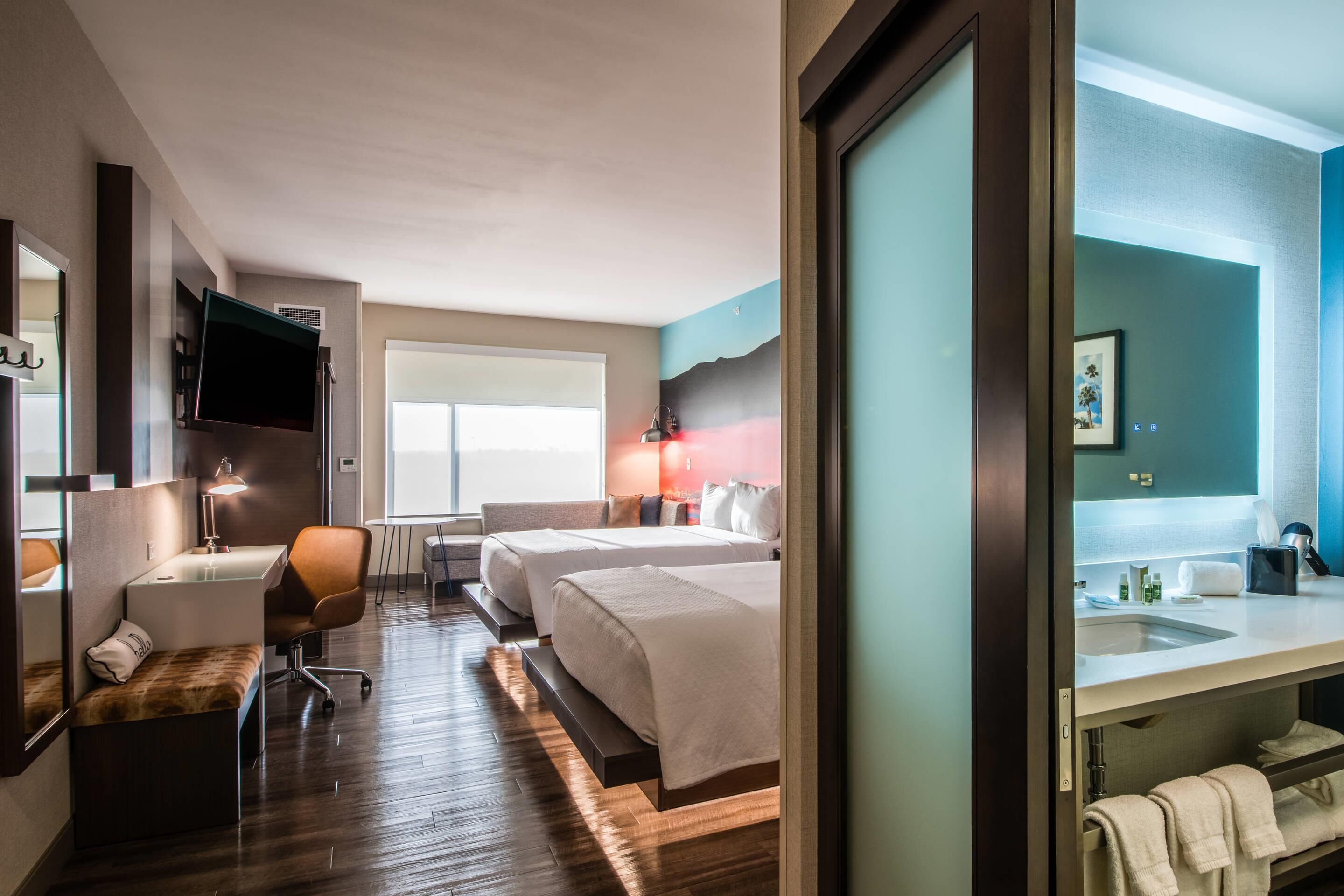 A hotel room with a bed, a desk, a chair, a TV, a window, and a bathroom with a sink and towel rack.