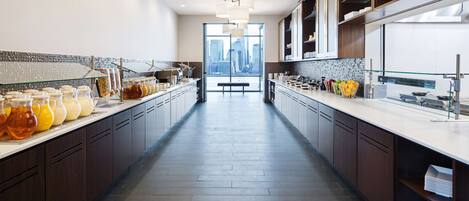 A modern kitchen with a long counter, glass containers of beverages, and a view of the city through large windows.