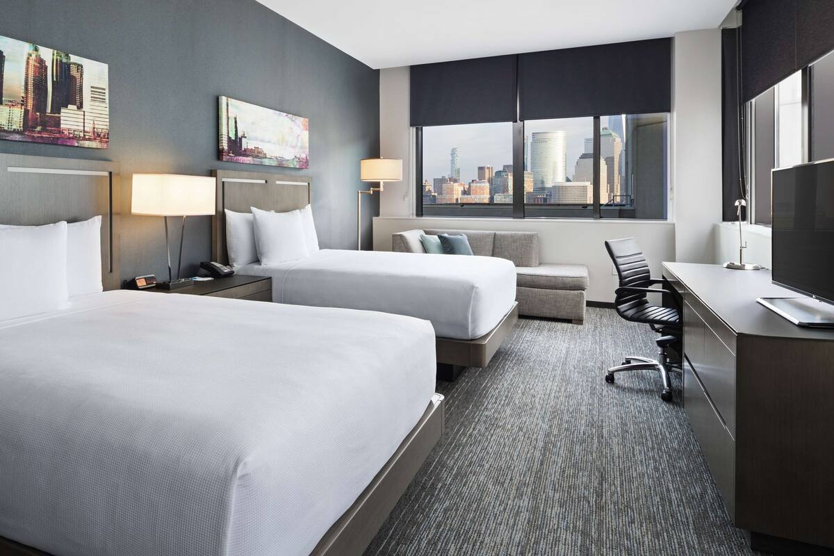 Une chambre d’hôtel avec deux lits, un bureau, une chaise, une lampe et une grande fenêtre donnant sur la ville.