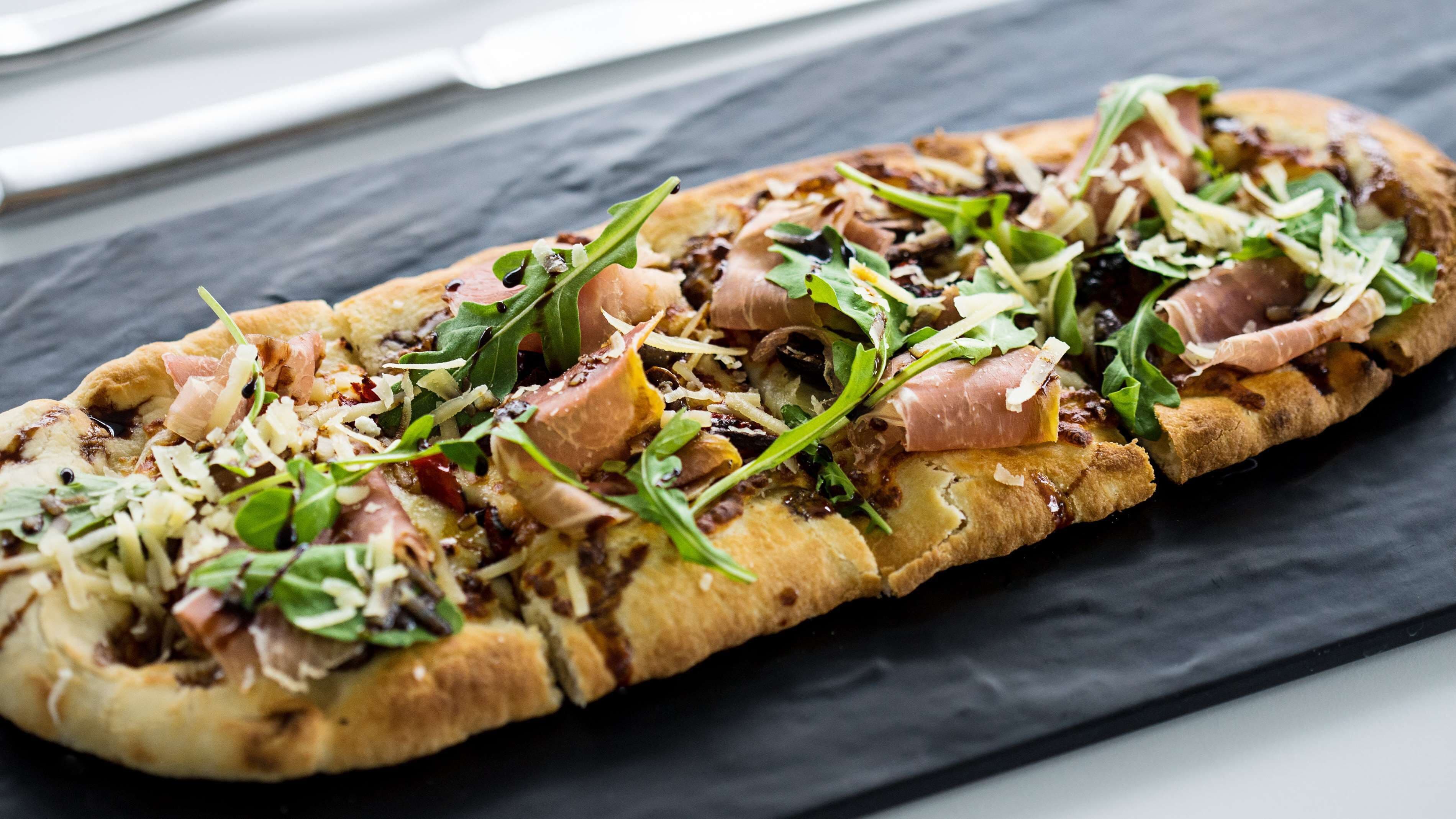 Una pizza larga y rectangular con varios ingredientes, incluyendo verduras, queso y carne, sobre una superficie de pizarra oscura.