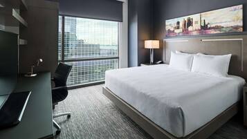 A hotel room with a large bed, a desk with a chair, a lamp, and a cityscape painting on the wall.