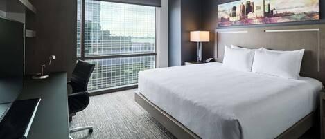 Habitación de hotel con una cama grande, un escritorio con silla, una lámpara y un cuadro de un paisaje urbano en la pared.