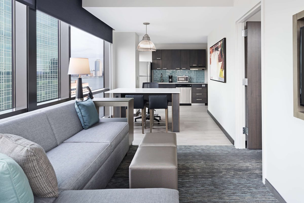 Un salon moderne avec un canapé gris, une petite table et un banc. Au fond, on aperçoit une cuisine avec des meubles sombres et un pare-feu en carrelage.