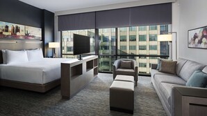 A hotel room with a large bed, a TV, a sofa, and a chair. There is a large window with a view of buildings.